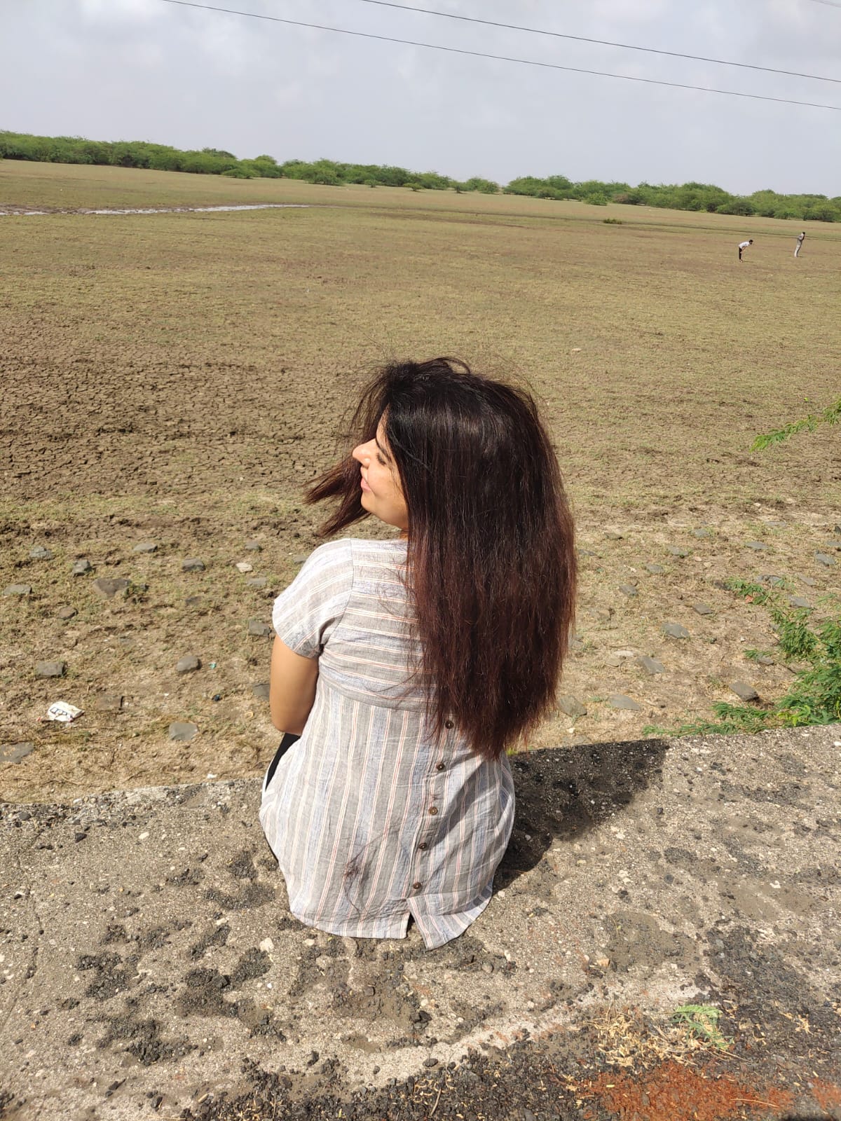 Woman sitting by a dry open field in a striped dress, enjoying a peaceful countryside view.