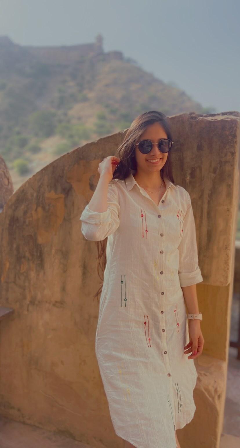 Woman in a white shirt dress and sunglasses posing outdoors with a scenic hill and fort backdrop.