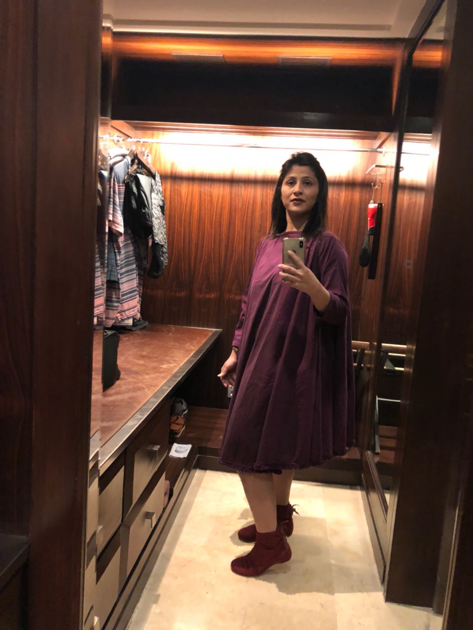 Woman taking a mirror selfie in a flowy purple dress and matching boots inside a stylish room.