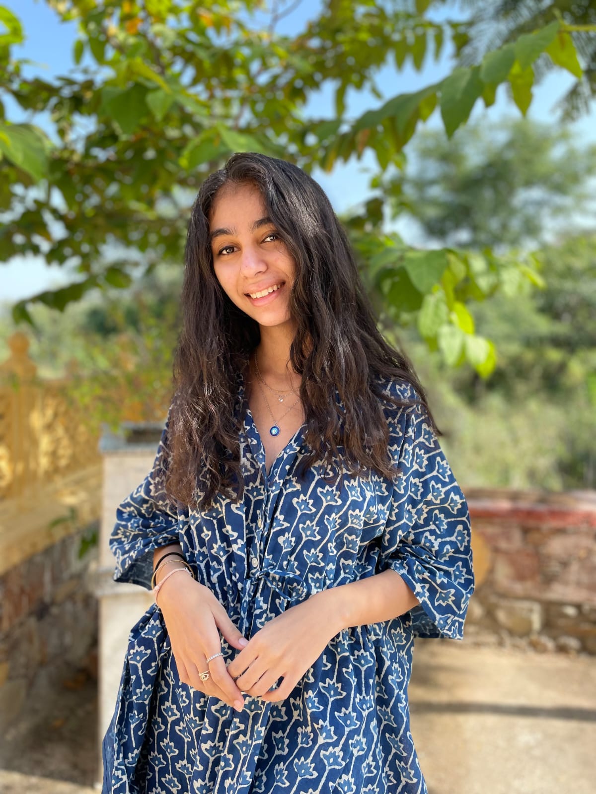 Woman in a blue printed dress smiling outdoors under trees in a natural sunlit setting.