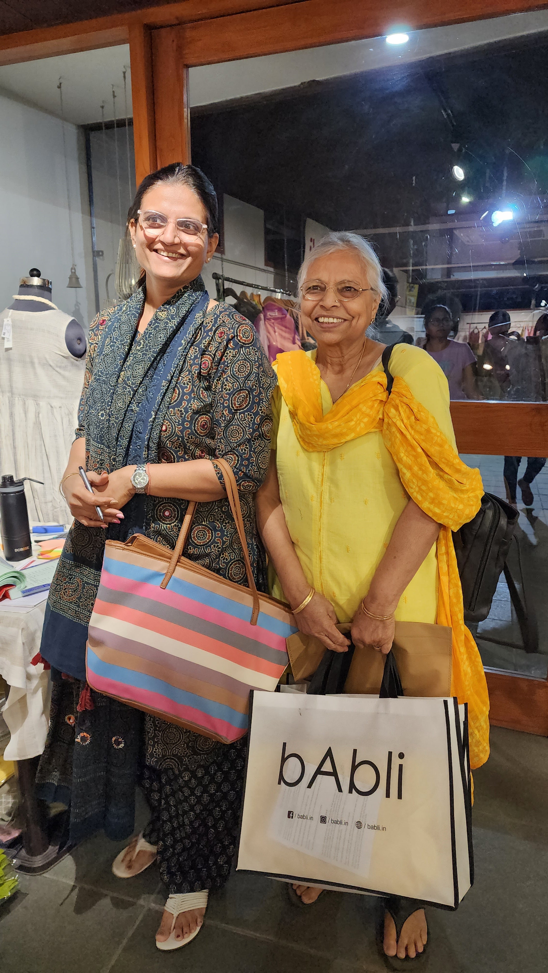 Two women smiling at a bAbli store, holding shopping bags and dressed in stylish ethnic wear.