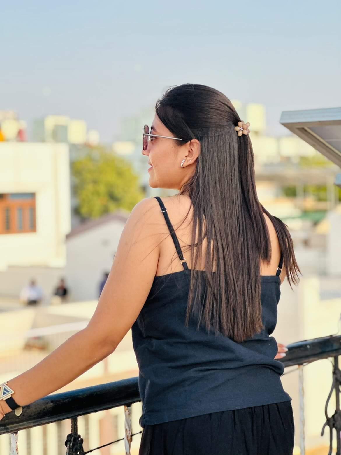 Woman in a black outfit standing on a balcony, enjoying a sunny city view with a relaxed pose.