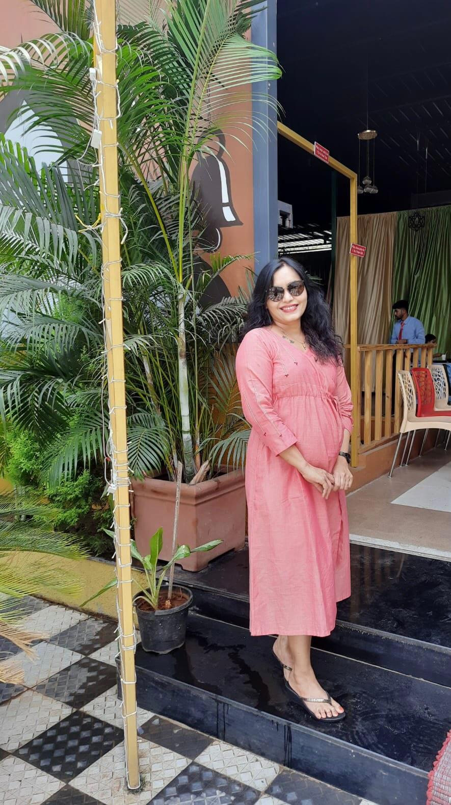 Woman in a pink dress and sunglasses posing outdoors near greenery and a cafe entrance.