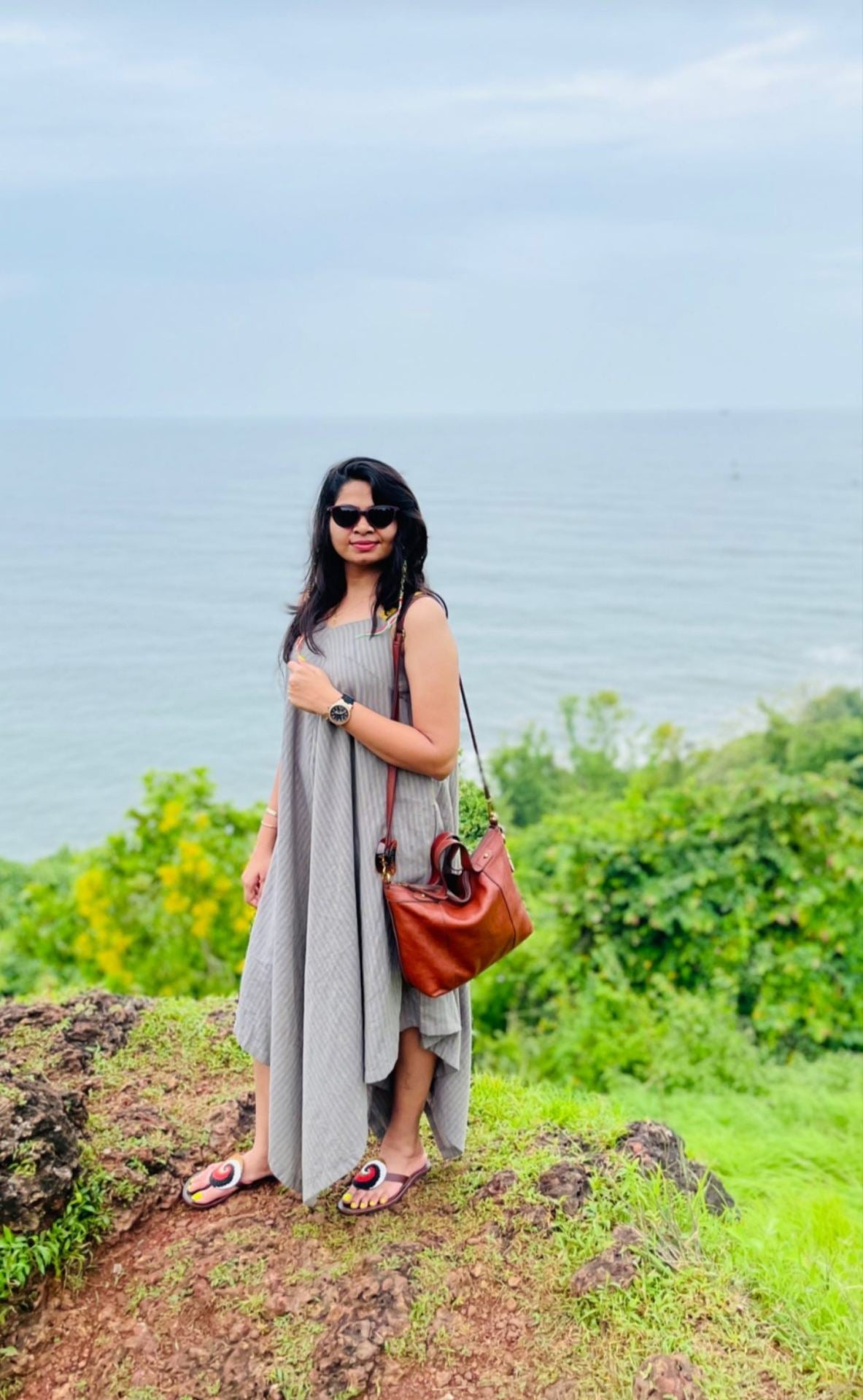 Woman in a grey dress and sunglasses posing on a hilltop with a scenic sea view in the background.