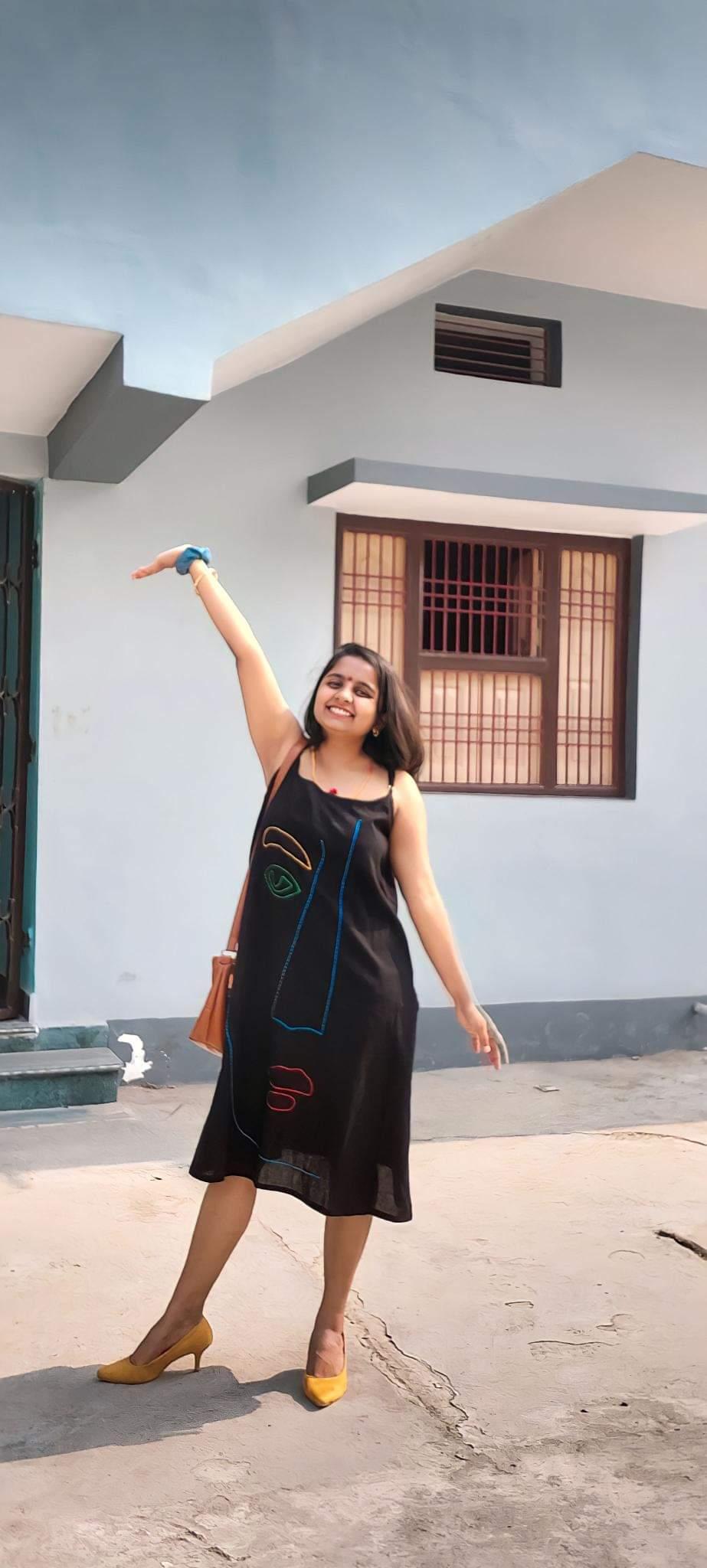 Woman in a black printed dress posing joyfully outdoors in front of a house on a sunny day.