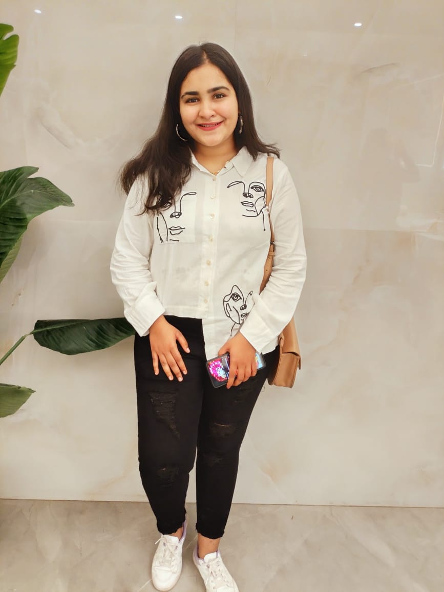 Woman in a white printed shirt and black jeans posing indoors with a casual and stylish look.